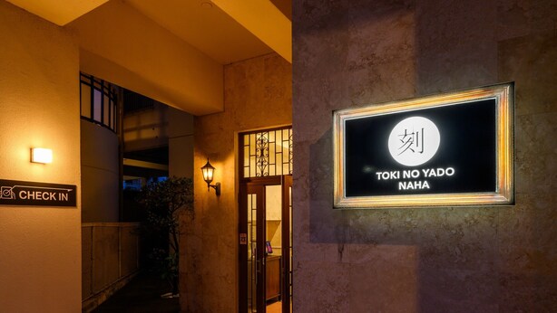 A hotel entrance with a 'Check In' sign, a lit wall-mounted sign with Japanese characters, and a door with a metal gate. - TOKI NO YADO NAHA by SUNREST RESORT (Naha)