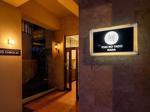 Entrada de un hotel con un letrero que dice 'Check In', un cartel iluminado con caracteres japoneses en la pared y una puerta con reja de metal.