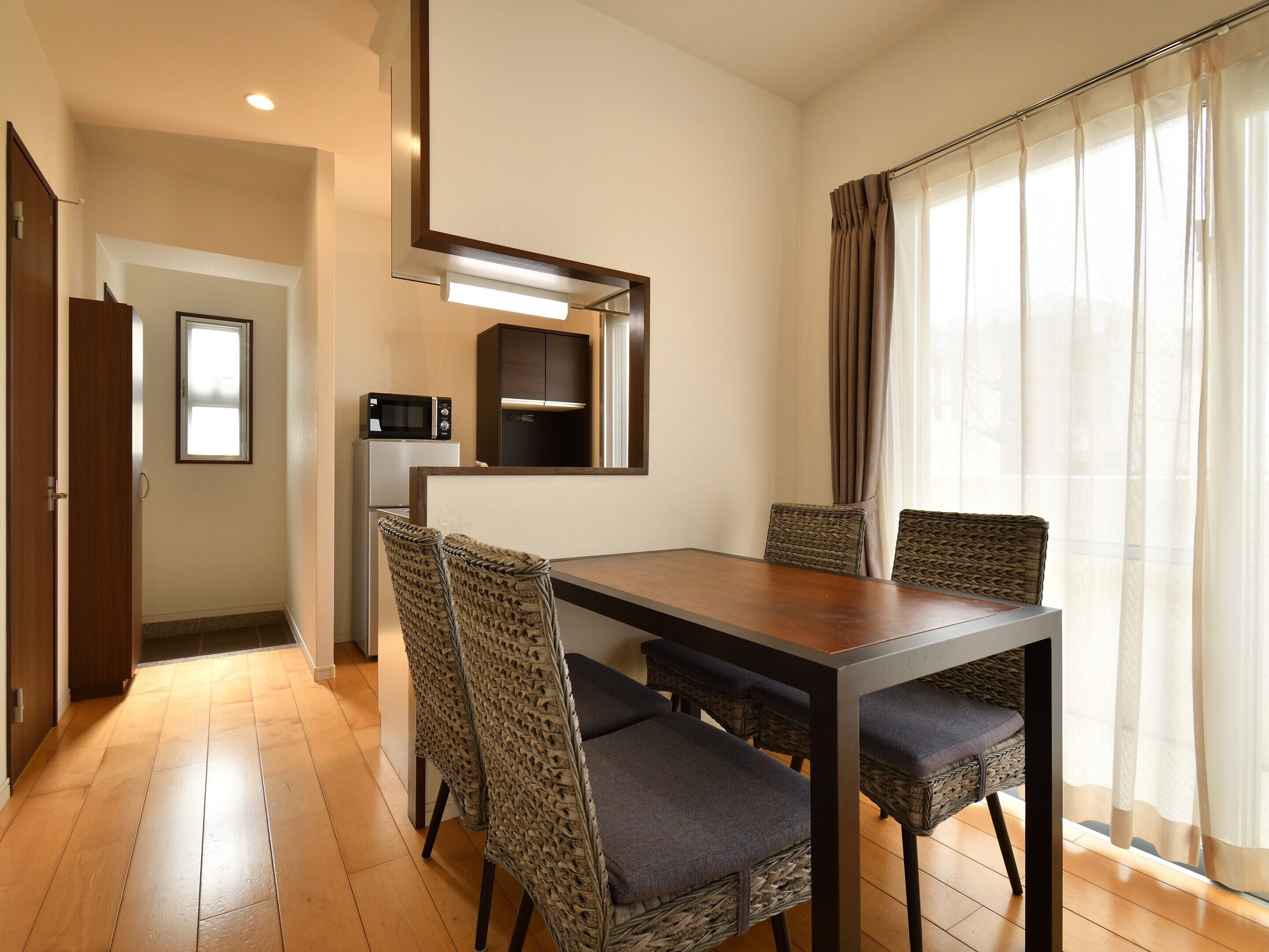 A dining area with a wooden table and woven chairs, a microwave, and a refrigerator in the background.