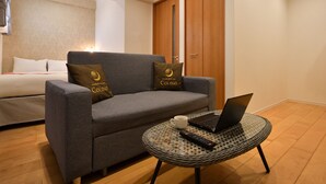 A modern hotel room with a grey sofa, a small round table with a laptop and remote control, a bed with white bedding, and a wooden wardrobe. - TOKI NO YADO NAHA by SUNREST RESORT (Naha)