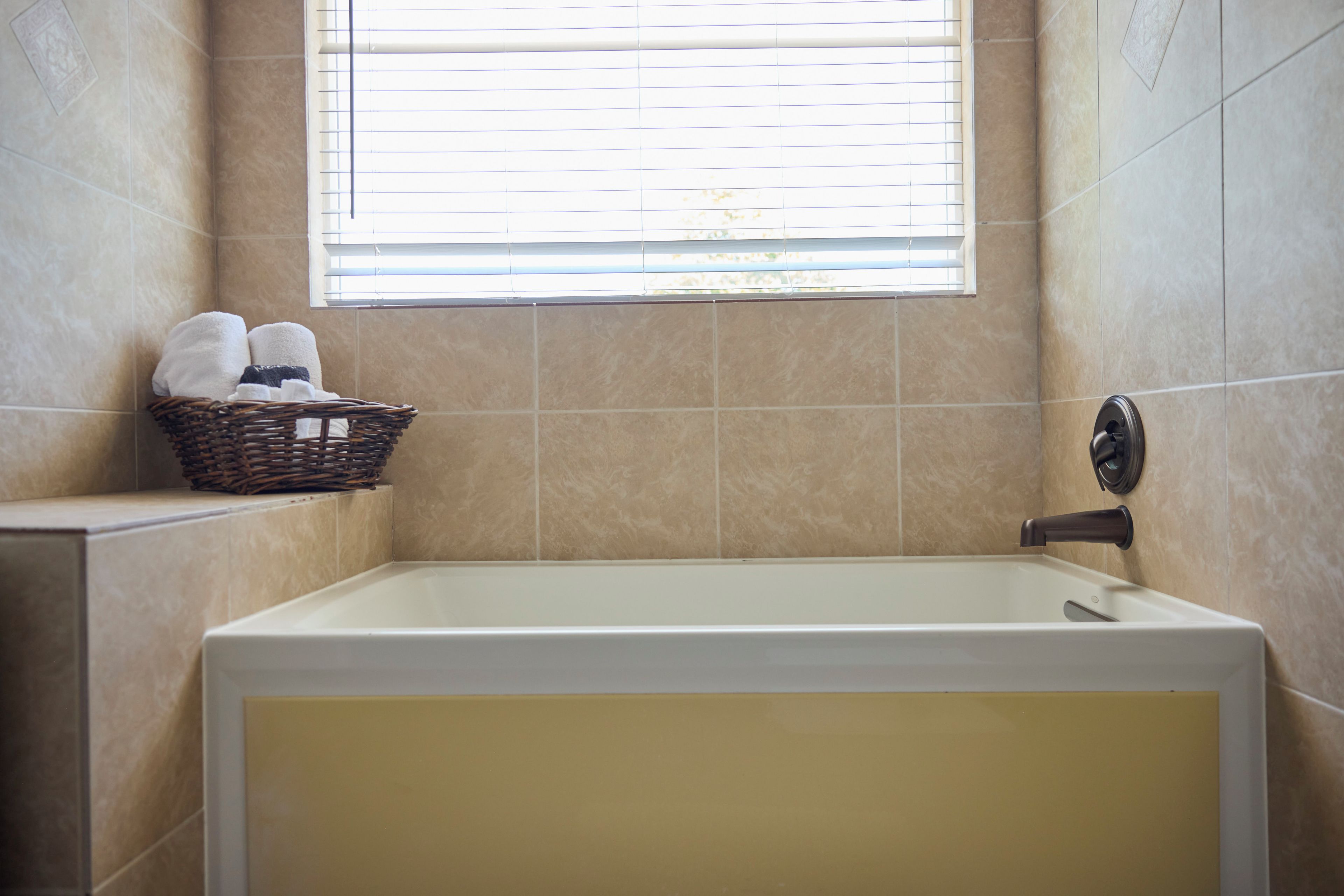 A bathroom with a bathtub, a window with blinds, and a wicker basket with towels.