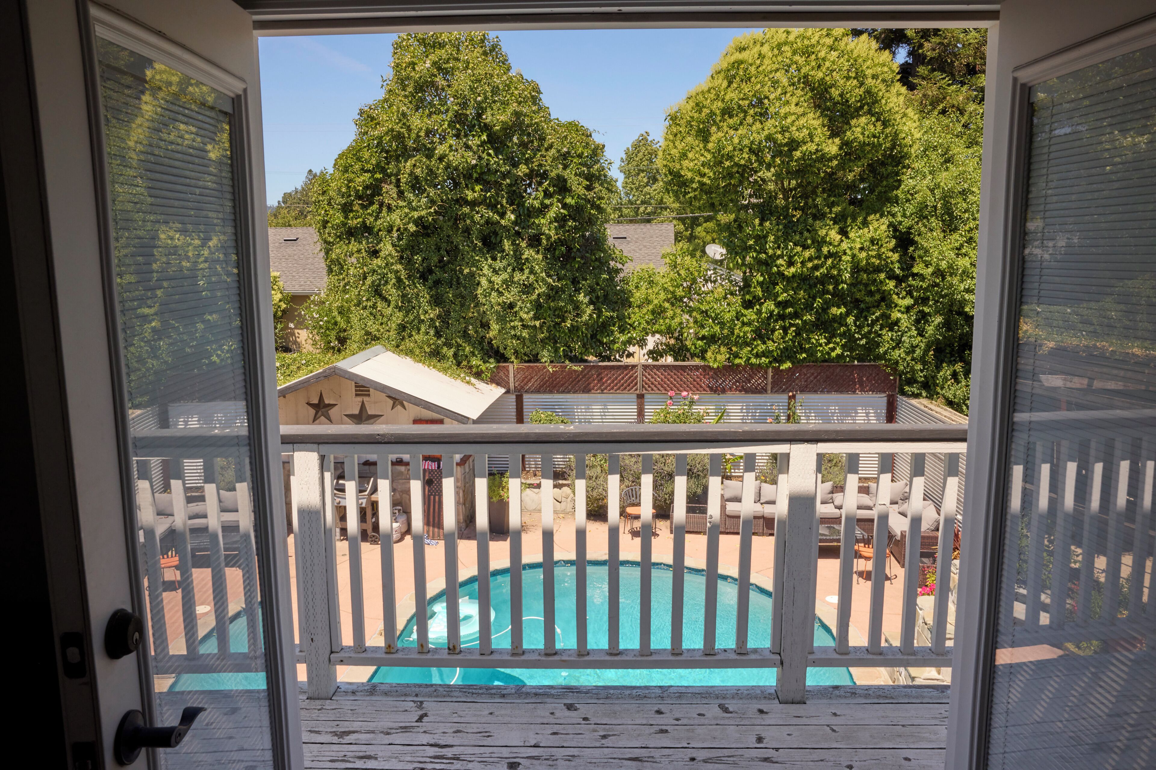 A view from a balcony with a swimming pool, surrounded by trees and a fenced area with outdoor furniture.