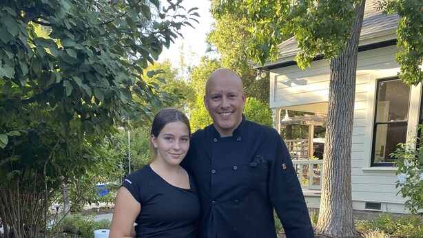 Un grand barbecue extérieur où sont préparées différentes sortes de viandes et de légumes, entouré d’un jardin et avec un bâtiment visible au loin.