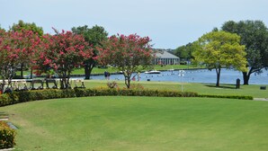 A golf course with a water body in the background, a building, and trees with pink flowers. - Beautiful 2/2 Apartment in Lake Conroe • Sleeps 6 (Montgomery)