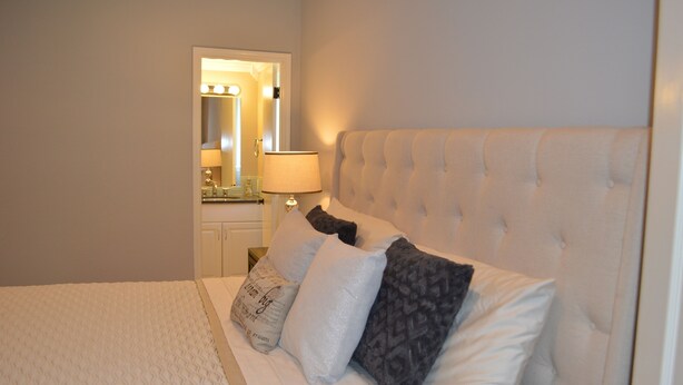 A neatly made bed with a tufted headboard, a bedside table with a lamp, and a mirror with lights above it. - Beautiful 2/2 Apartment in Lake Conroe • Sleeps 6 (Montgomery)