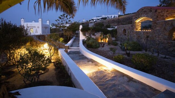 A stone pathway leading to a white building with a red roof, surrounded by greenery and palm trees. - Villa Hurmuses (Mykonos)