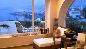 A room with a large window overlooking a coastal view, a bed, a chair, a small table with books and a bowl of fruit. - Villa Hurmuses (Mykonos)