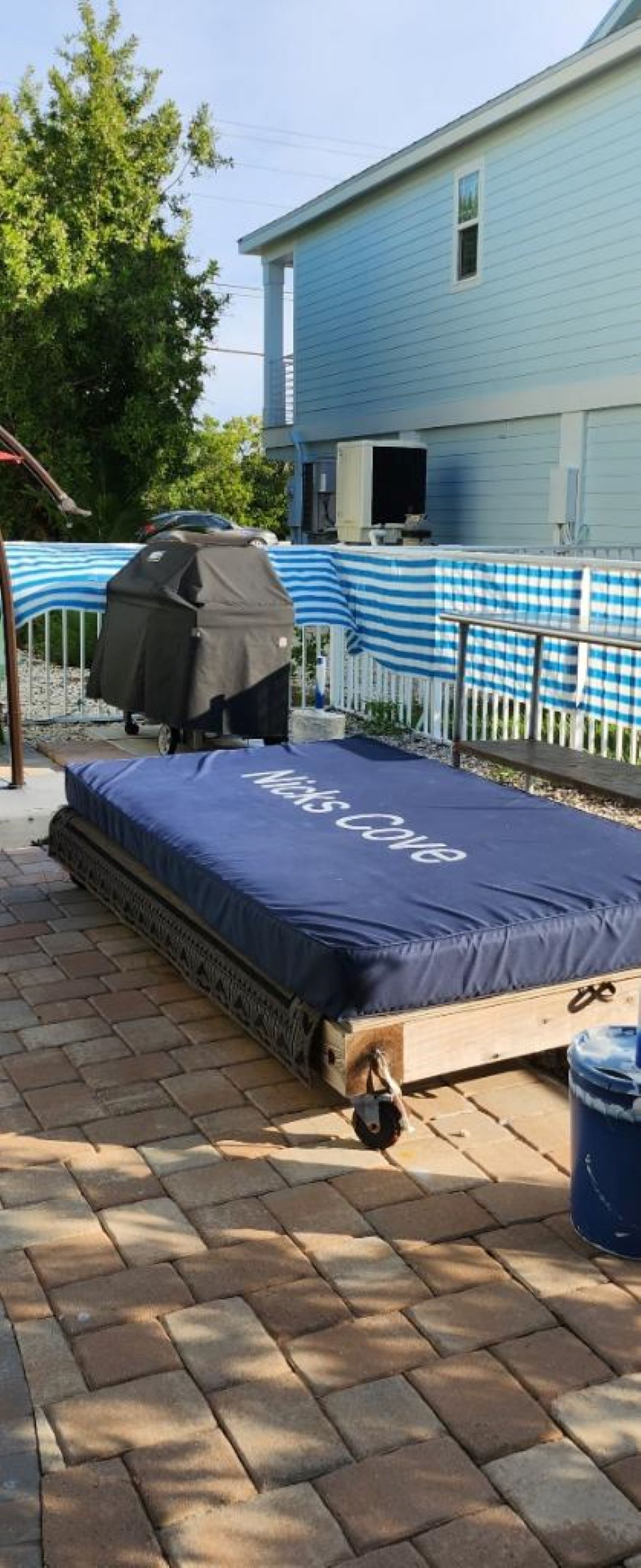 A mattress on a wooden platform with a grill and a bucket, a blue house in the background, and a fenced area with blue and white checkered fabric.