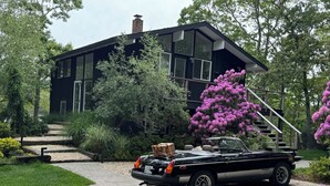 Uma casa moderna preta com entrada por carro, rodeada por vegetação exuberante e um carro conversível estacionado.