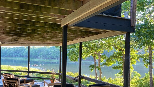 Um deck de madeira com espreguiçadeiras e uma mesa, com vista para um lago cercado por árvores e um píer.