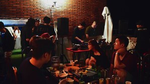 Eine Gruppe von Personen ist um einen Tisch mit Essen und Getränken versammelt, in einem Raum mit Backsteinwänden und einem grünen Teppich.