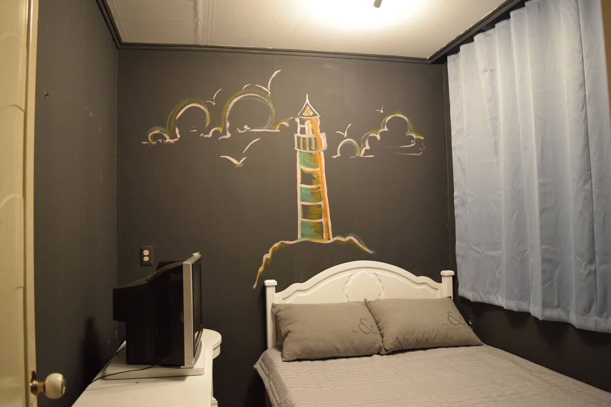 A bedroom with a bed, a TV on a stand, and a wall mural of a lighthouse.