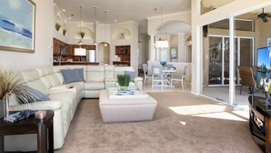 Un salon spacieux avec un canapé en cuir blanc, un pouf central et un coin repas à l’arrière-plan.