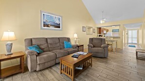 A living room with a sofa, armchair, wooden coffee table, and lamps. There is a painting on the wall and a kitchen area in the background. - 2/2 Vaulted Ceiling 10 Min to beach on golf course (Venice)