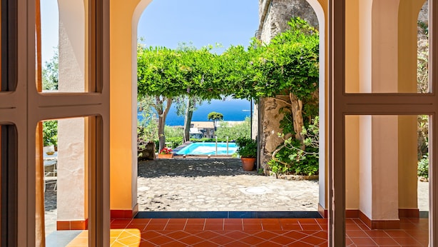 Puerta abierta que da a un patio con piscina, rodeado de vegetación y vistas al mar.