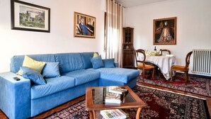 Una sala de estar con un sofá azul, una mesa de centro de vidrio con libros, un mueble de madera y dos cuadros en la pared.