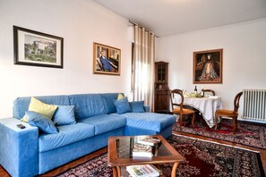 A living room with a blue sofa, a glass coffee table with books, a wooden cabinet, and two framed paintings on the wall.
