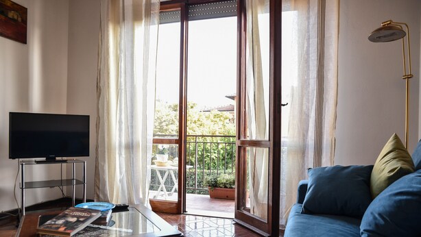 Una sala de estar con televisor, una mesa de centro con libros, un sofá azul y vistas a un balcón con plantas.
