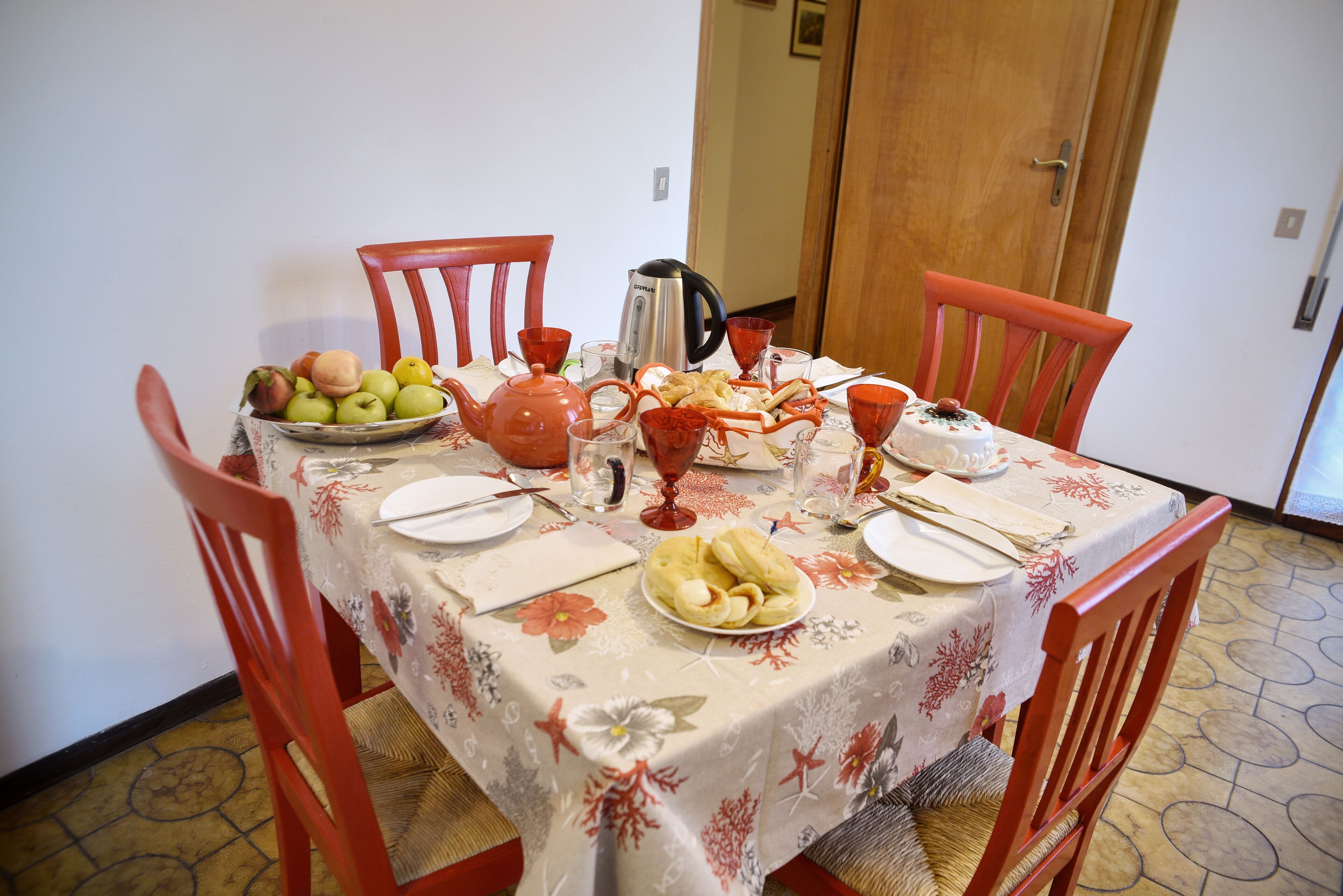 Une table à manger dressée avec une nappe à motifs floraux, des chaises rouges, une bouilloire, un panier de fruits et une assiette de raviolis.