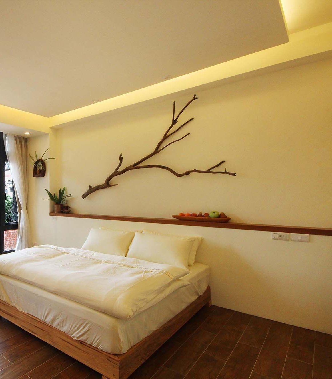 A bedroom with a bed, a branch wall art piece, a shelf with plants, and a window with curtains.