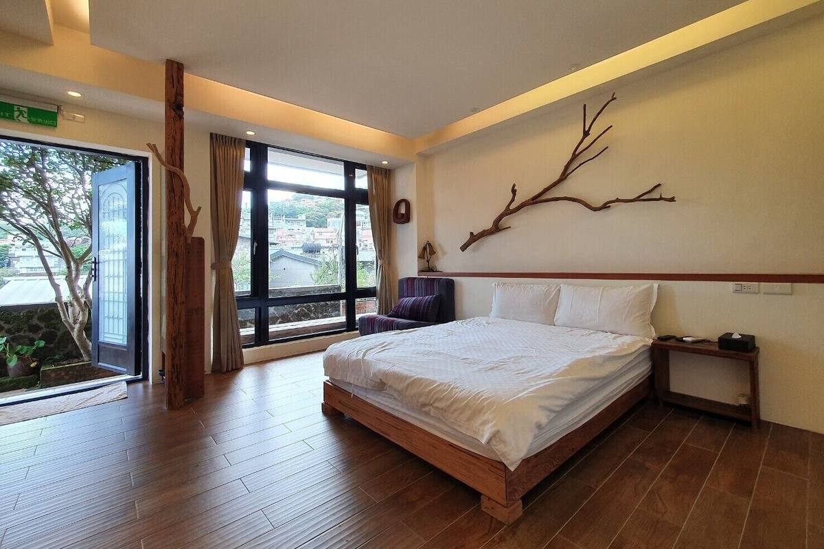 Quarto moderno com uma cama grande, piso de madeira, vista através de uma porta de correr de vidro e um enfeite de galho na parede.