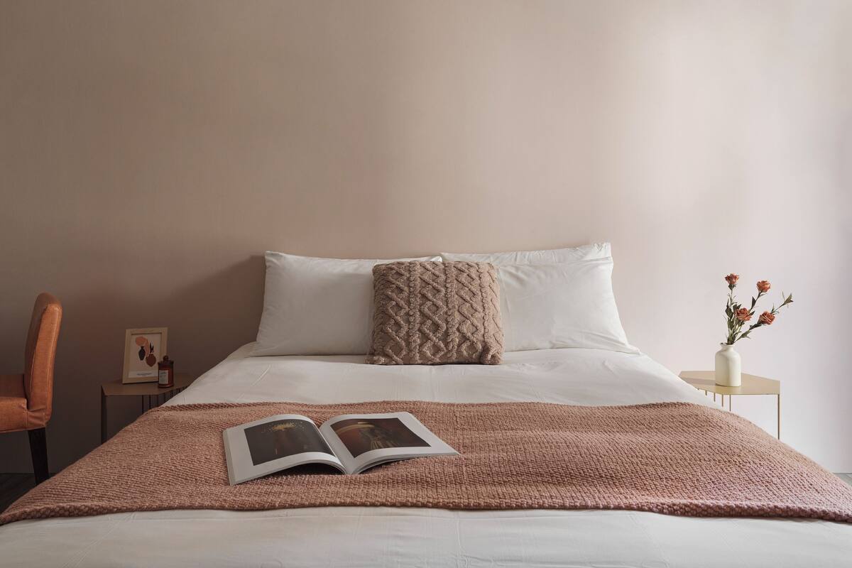 Una cama bien hecha con un libro, un cojín decorativo y un florero con flores sobre una mesita de noche.