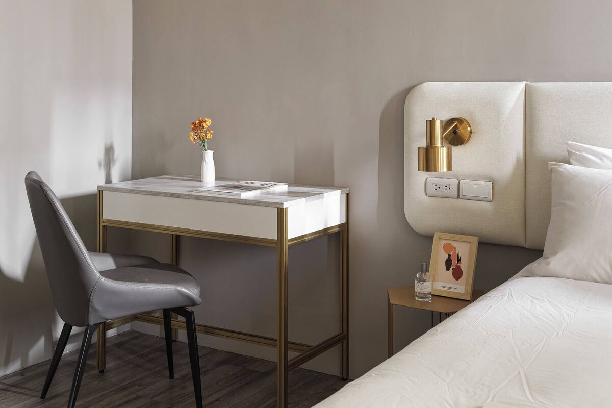 Habitación de hotel moderna con diseño minimalista, que incluye un escritorio, una silla cómoda, una cama con ropa blanca y una mesita de noche con un cuadro enmarcado.