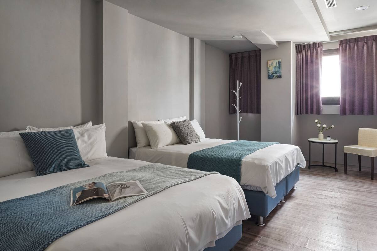 Habitación de hotel con dos camas, una mesita con un florero y una ventana con cortinas.