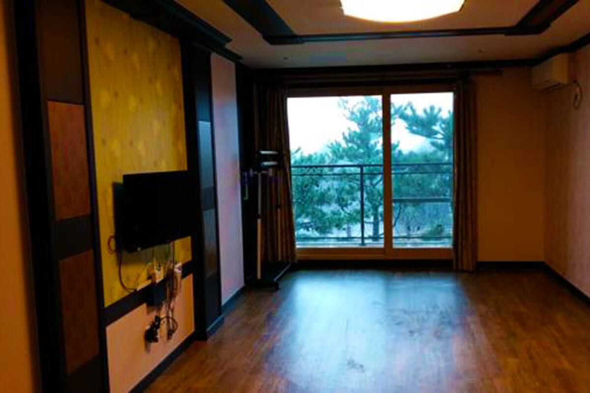 Een kamer met een grote schuifglazen deur die leidt naar een balkon, houten vloeren en een wandgemonteerde flatscreen televisie aan de linkerwand.