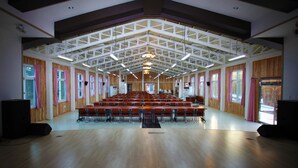 Een ruime zaal met rijen stoelen, houten wanden en een hoog plafond met zichtbare balken.