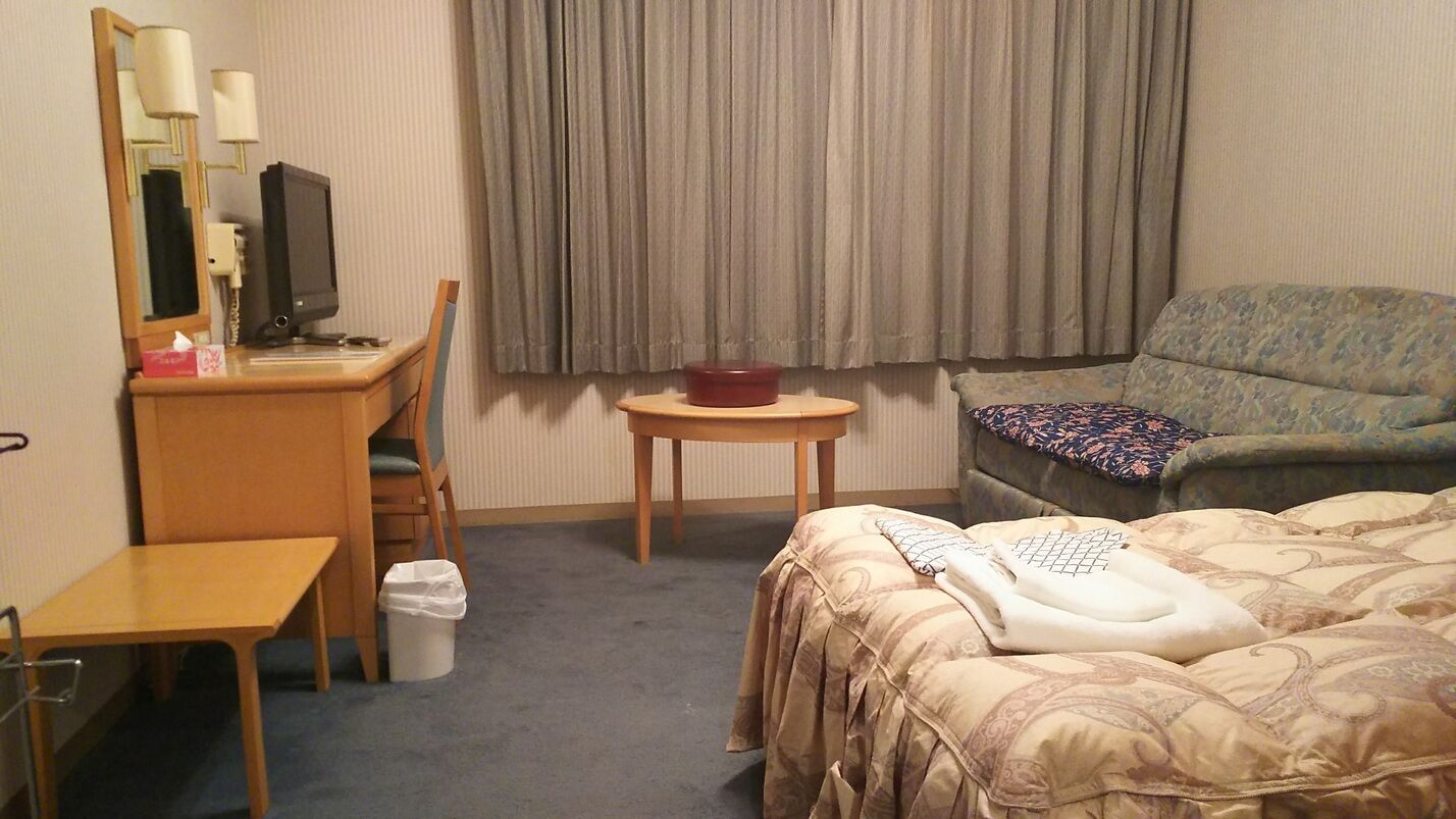 A hotel room with a bed, a desk, a chair, a television, a small table, and a window with curtains.