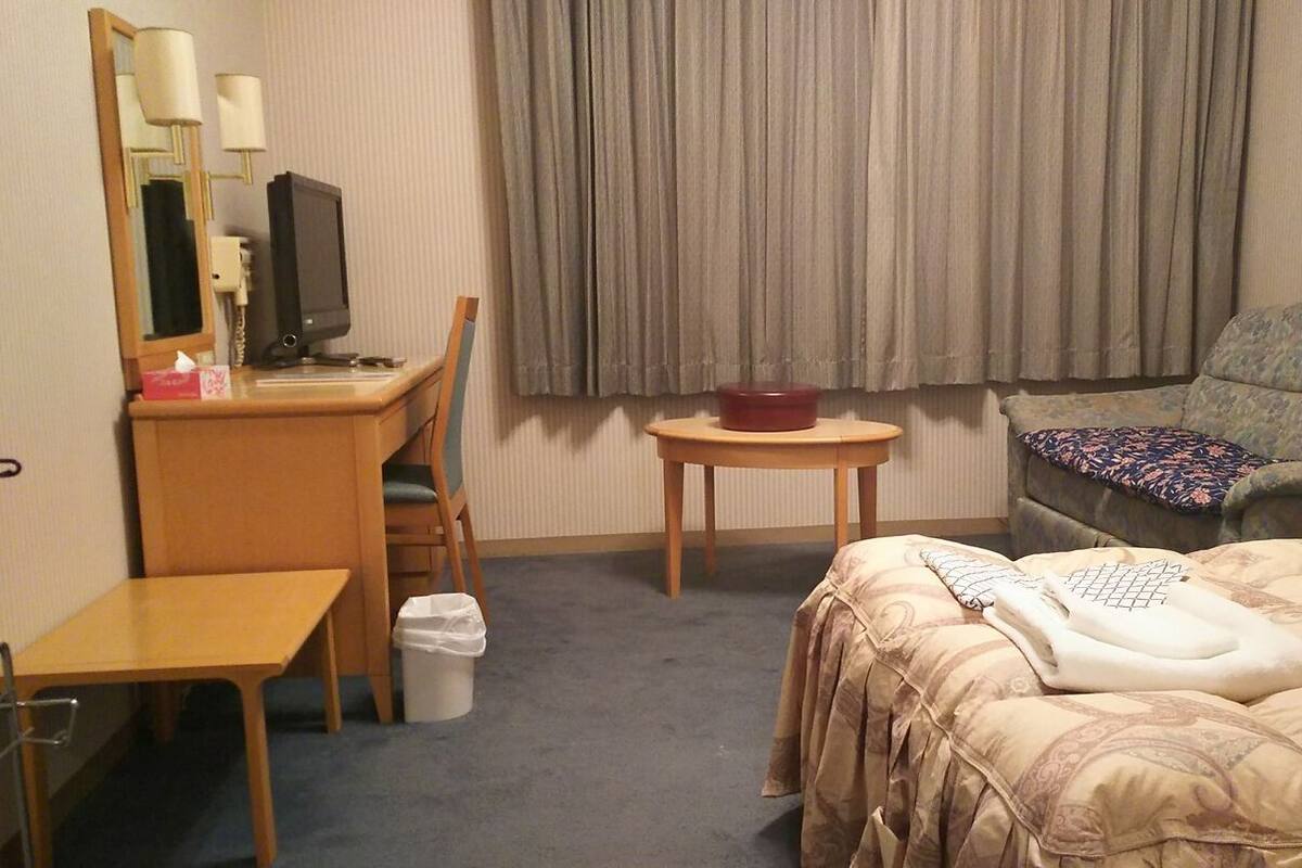 Ein Hotelzimmer mit Bett, Schreibtisch, Stuhl, Fernseher, kleinem Tisch und einem Fenster mit Vorhängen.