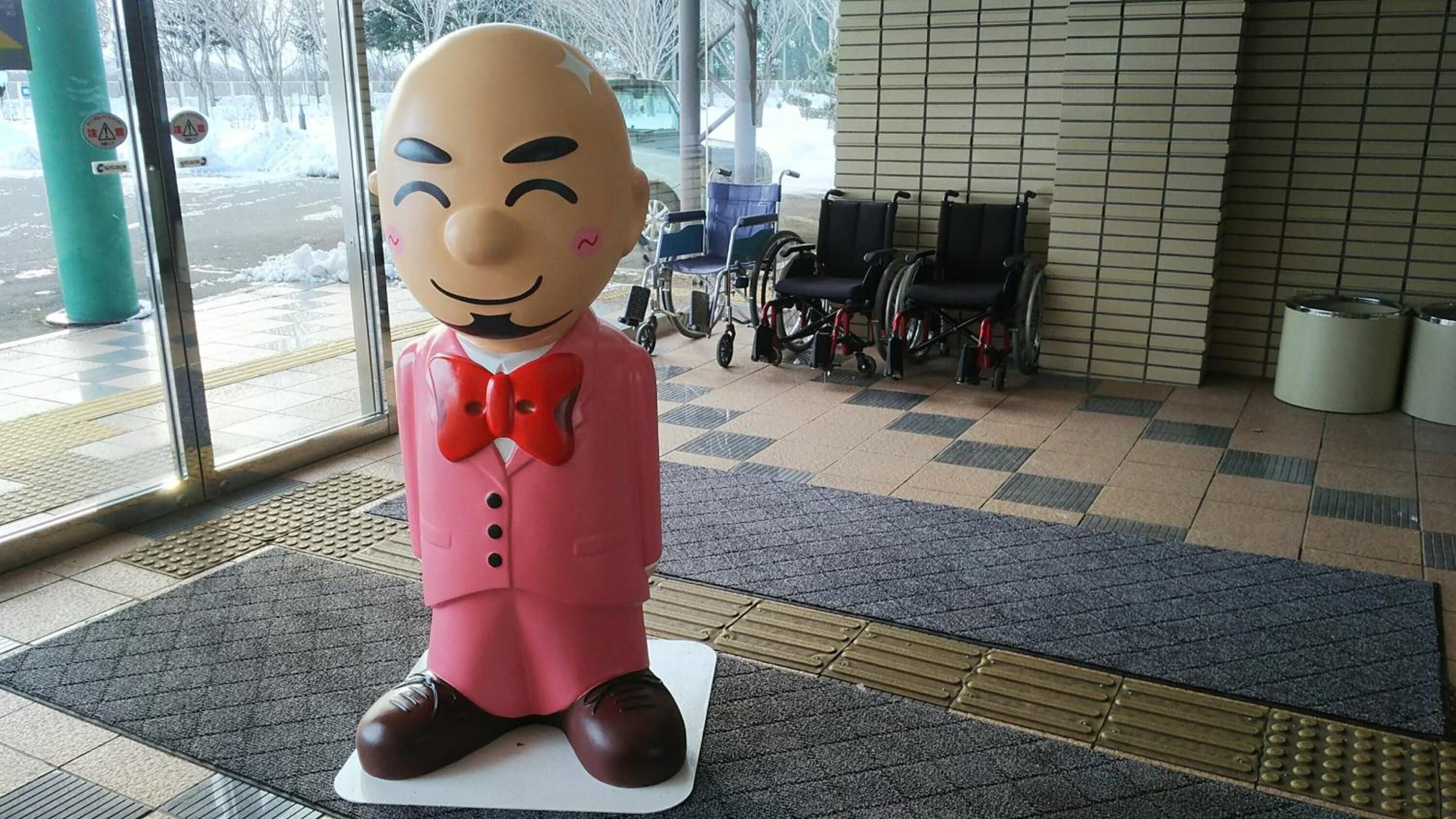 A large statue of a character in a pink suit and red bow tie, wheelchair-accessible seating, and a tiled floor with tactile paving.
