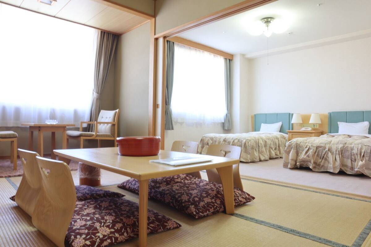 Ein Hotelzimmer mit Tatami-Boden, einem Holztisch mit roter Schüssel, zwei Betten mit blauen Kopfteilen und einem Fenster mit Vorhängen.