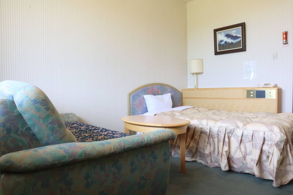 Ein Hotelzimmer mit Bett, Sessel, Nachttisch, Lampe und einem gerahmten Bild an der Wand.