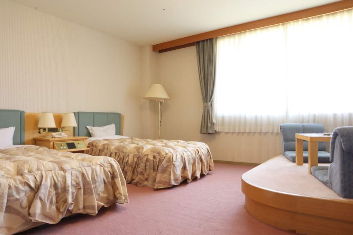 Ein Hotelzimmer mit zwei Betten, einem Schreibtisch, einem Stuhl, einer Lampe und einem Fenster mit Vorhängen.