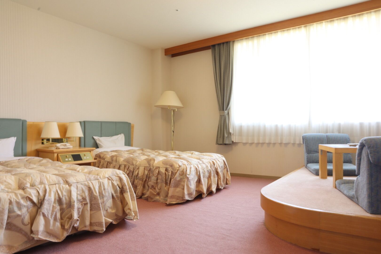 A hotel room with two beds, a desk, a chair, a lamp, and a window with curtains.