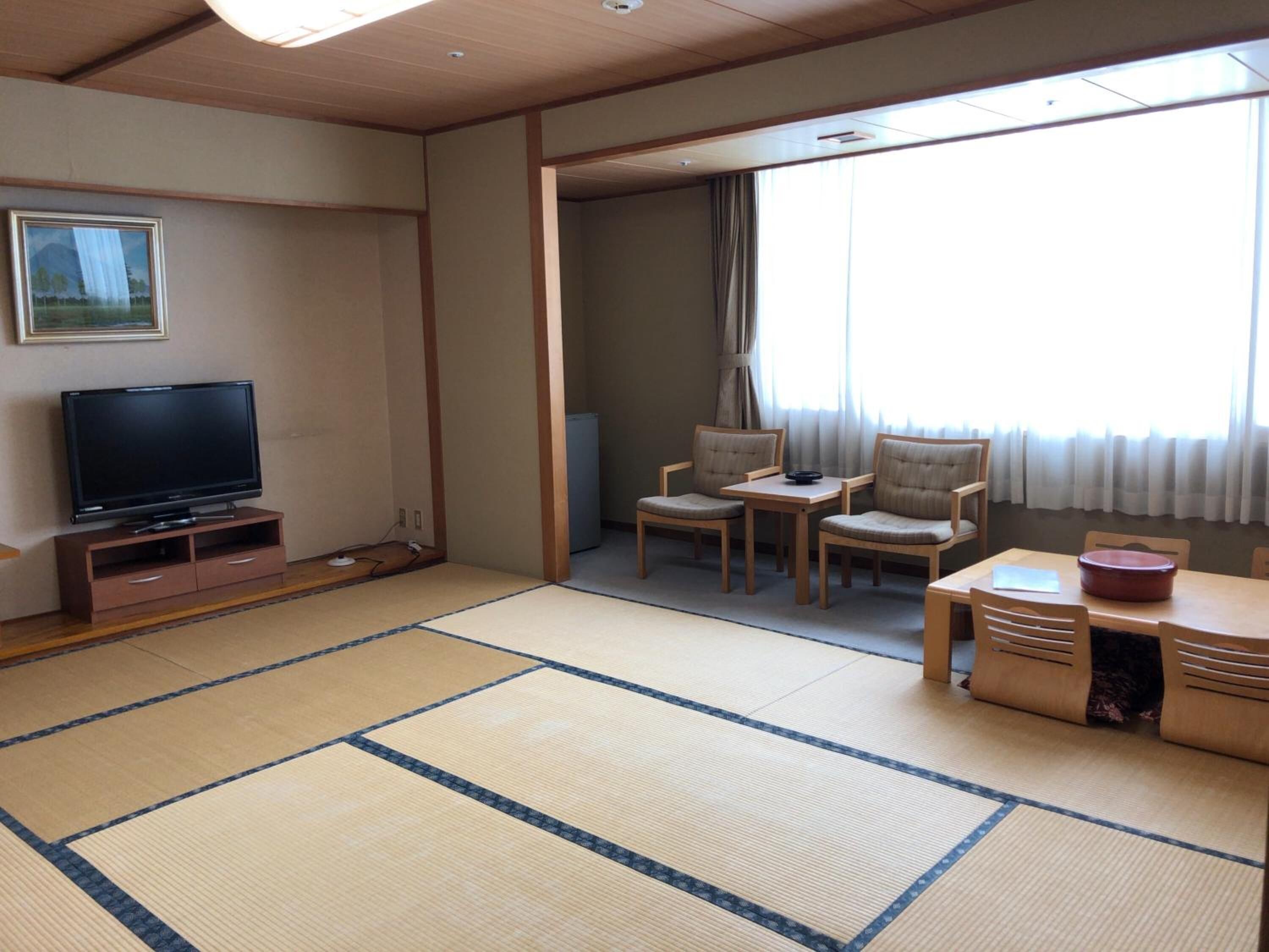 Habitación con esteras de tatami, televisión, mesa de comedor con sillas y vistas al exterior.