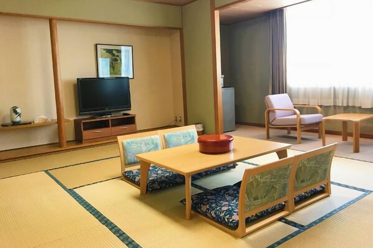 Een traditionele Japanse kamer met tatami vloerbedekking, een televisie, een eettafel met kussens en een bank.