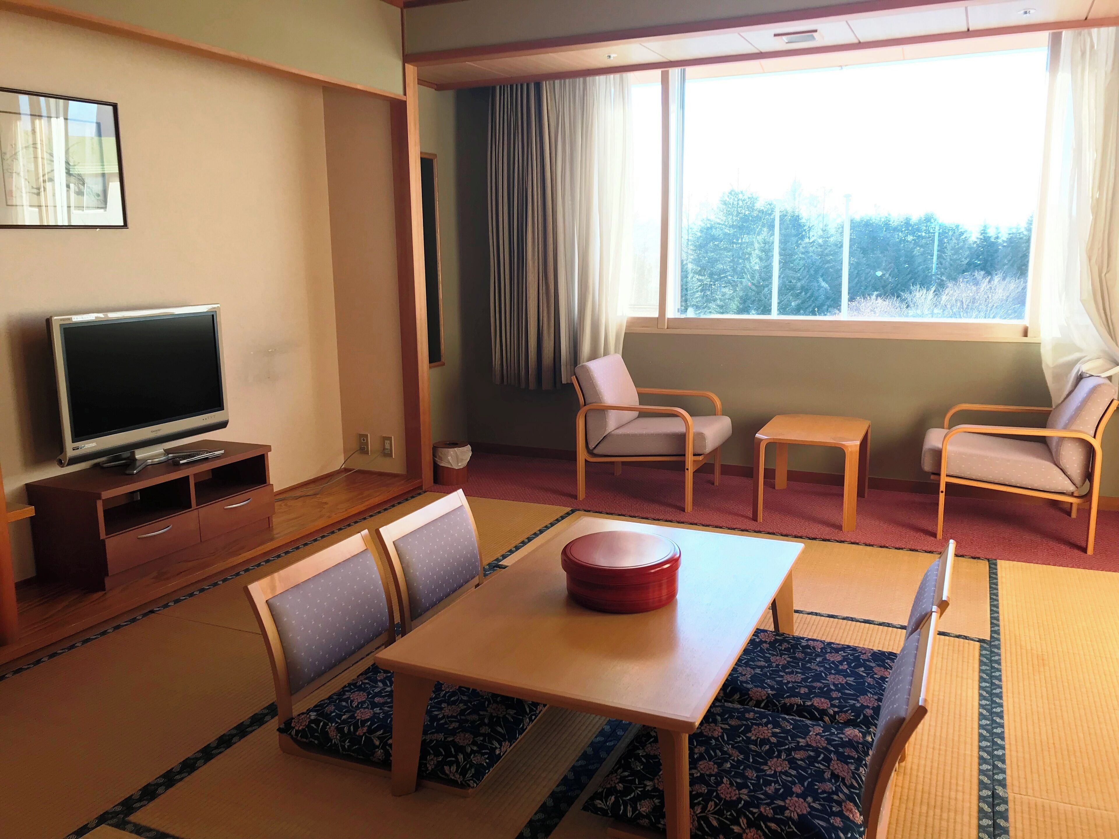 Una habitación con una televisión sobre un soporte de madera, una mesa de comedor con sillas y una ventana con cortinas.