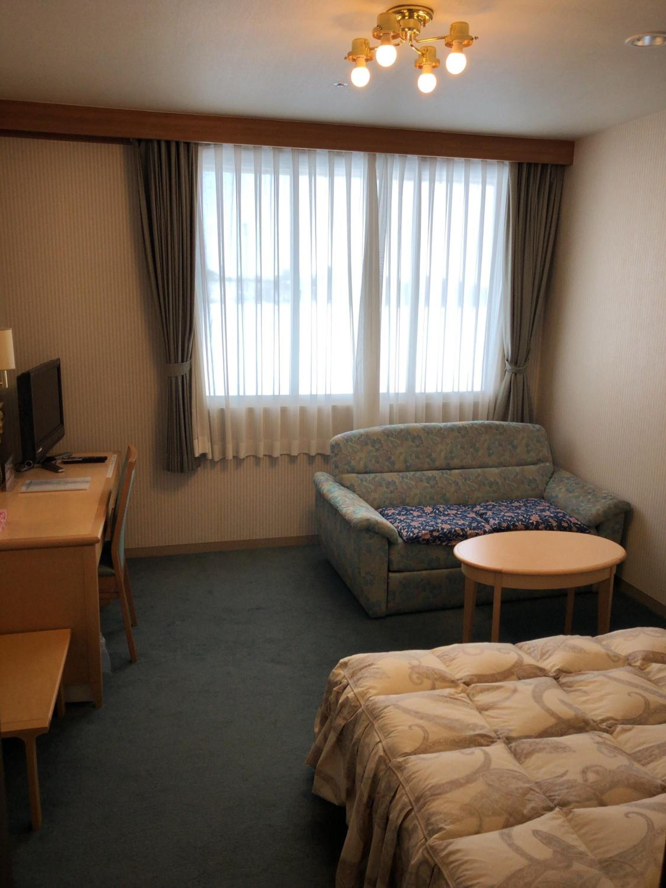 Hotellihuone, jossa on sänky, sohva, pieni pöytä, työpöytä tietokoneella ja ikkuna, jossa on verhot.