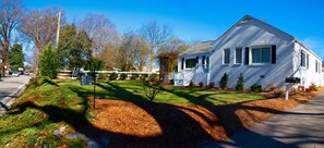A white two-story house with a front porch, a mailbox, and a well-maintained lawn with mulch and small shrubs.