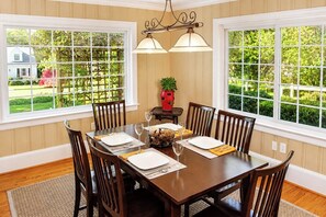 A dining room with a wooden table set for four, large windows with a view of greenery, and a chandelier.