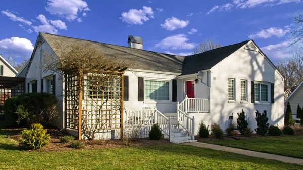 A two-story white house with a red door, a covered porch, and a small front yard with a wooden fence and shrubs.