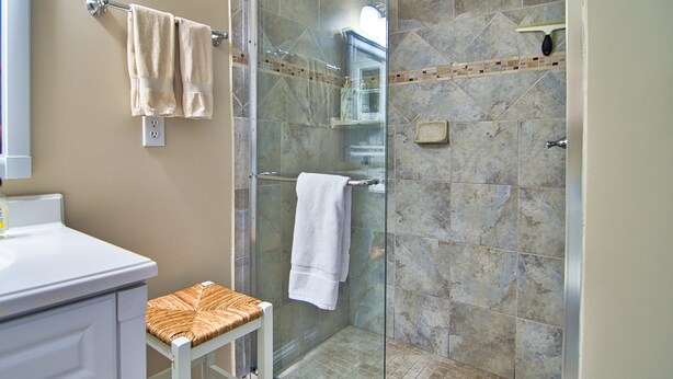 A bathroom with a glass shower enclosure, a white sink, a white cabinet, and a wicker stool.