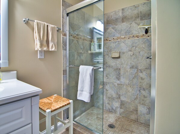 A bathroom with a glass shower enclosure, a white sink, a white cabinet, and a wicker stool.