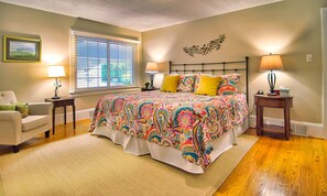 A bedroom with a large bed, two bedside tables, a chair, a window with blinds, and a decorative wall piece.