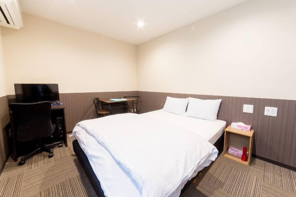 A hotel room with a bed, a desk with a chair, a TV, and a small bedside table with a vase.