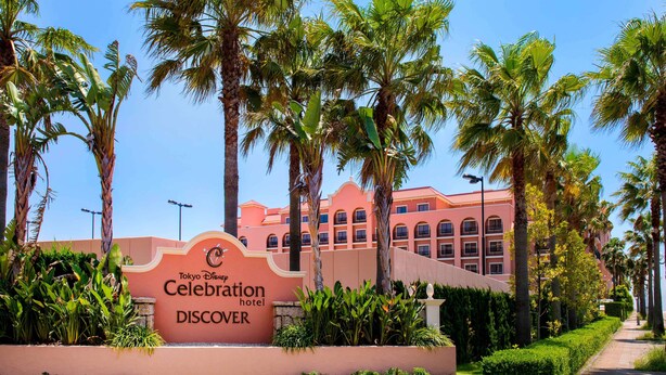 A sign for the Tokyo Disney Resort Celebration Hotel Discover with palm trees and a building in the background.