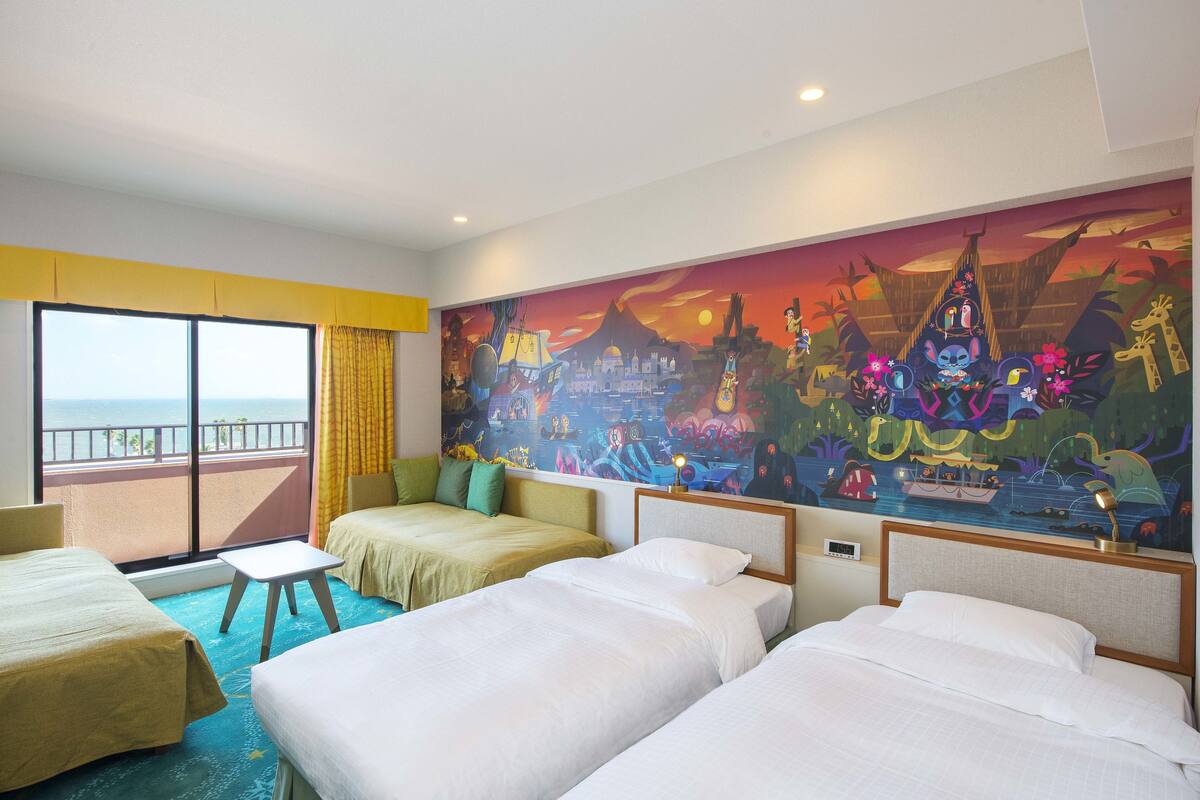 A hotel room with three beds, a small table, and a large window with a view of the sea.